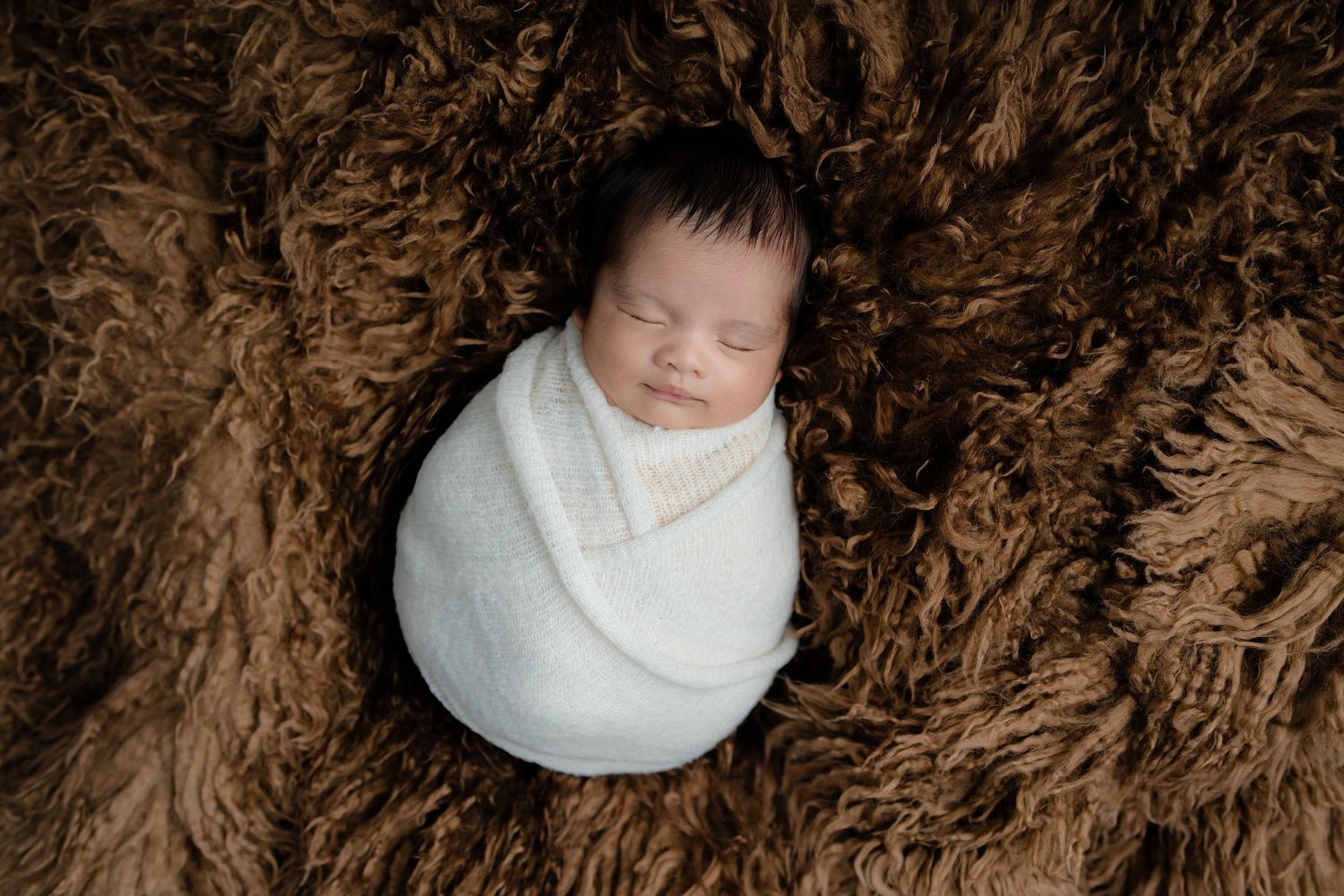 A sleeping newborn wearing a white wrap lies peacefully on a brown textured blanket.