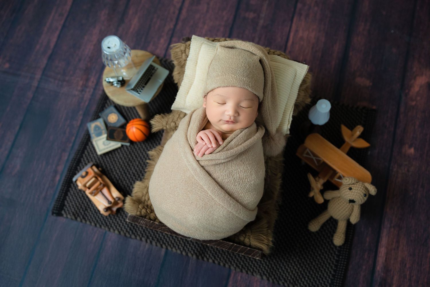 Sleeping newborn swaddled in beige wrap on dark wood floor with vintage toys and decorative items.