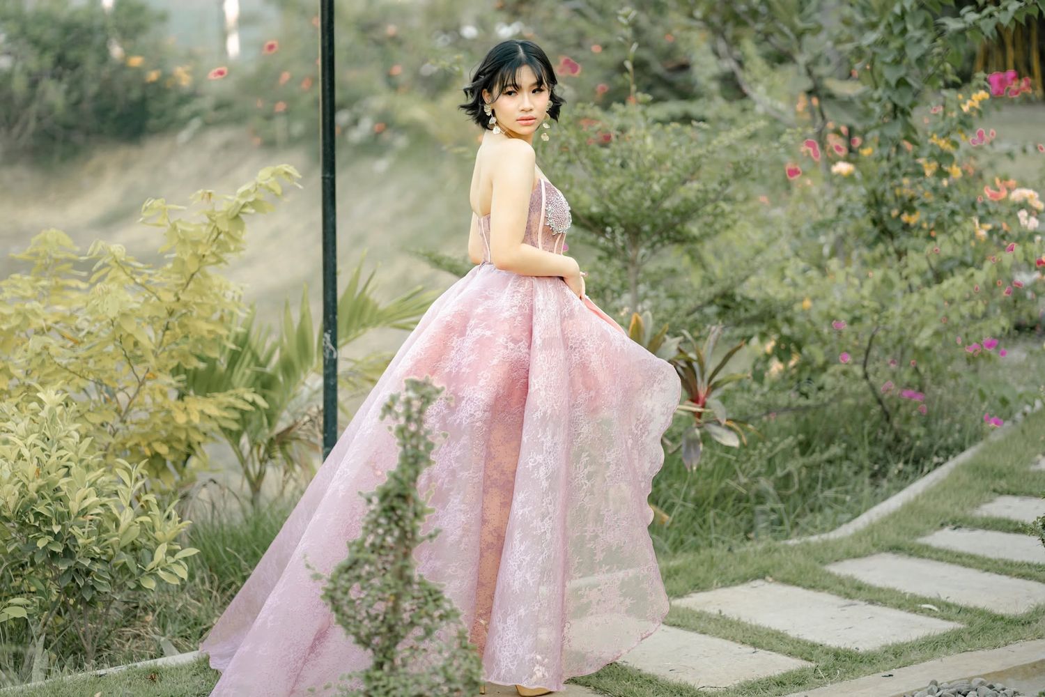 Young woman in elegant pink floral ball gown poses gracefully in a lush garden setting.