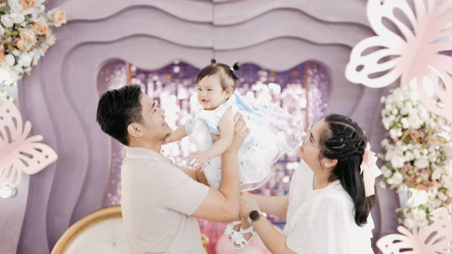 A joyful family celebrates their baby's milestone with purple butterfly decorations and floral backdrop.