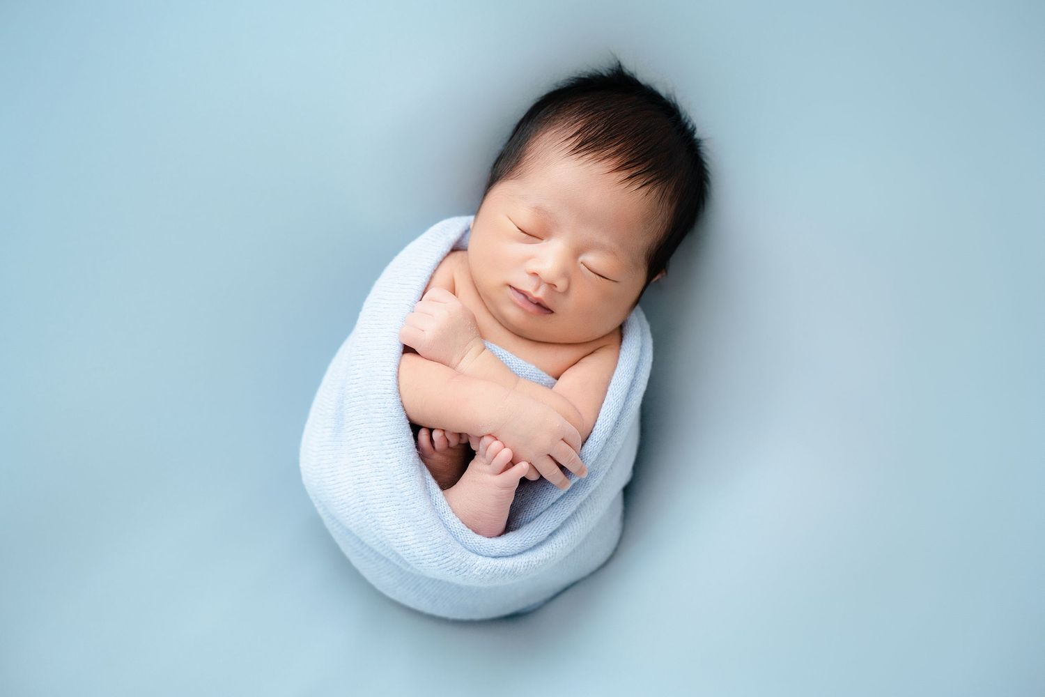 Peaceful newborn wrapped in white fabric against light blue background for professional portrait.