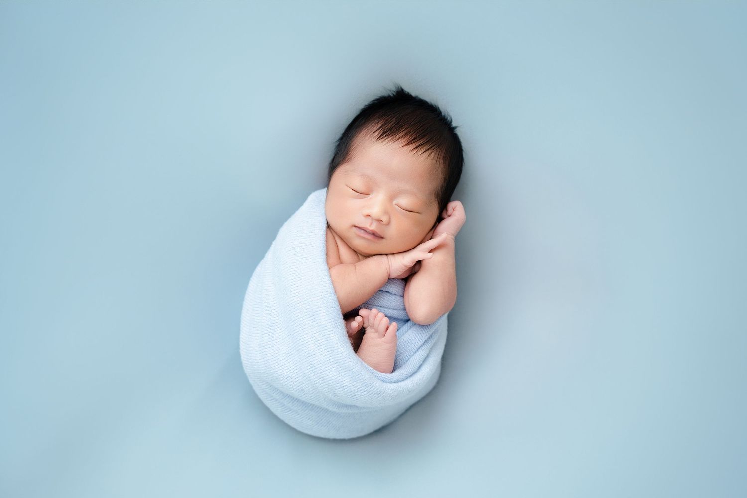 Professional newborn portrait with baby swaddled in white against soft blue background.