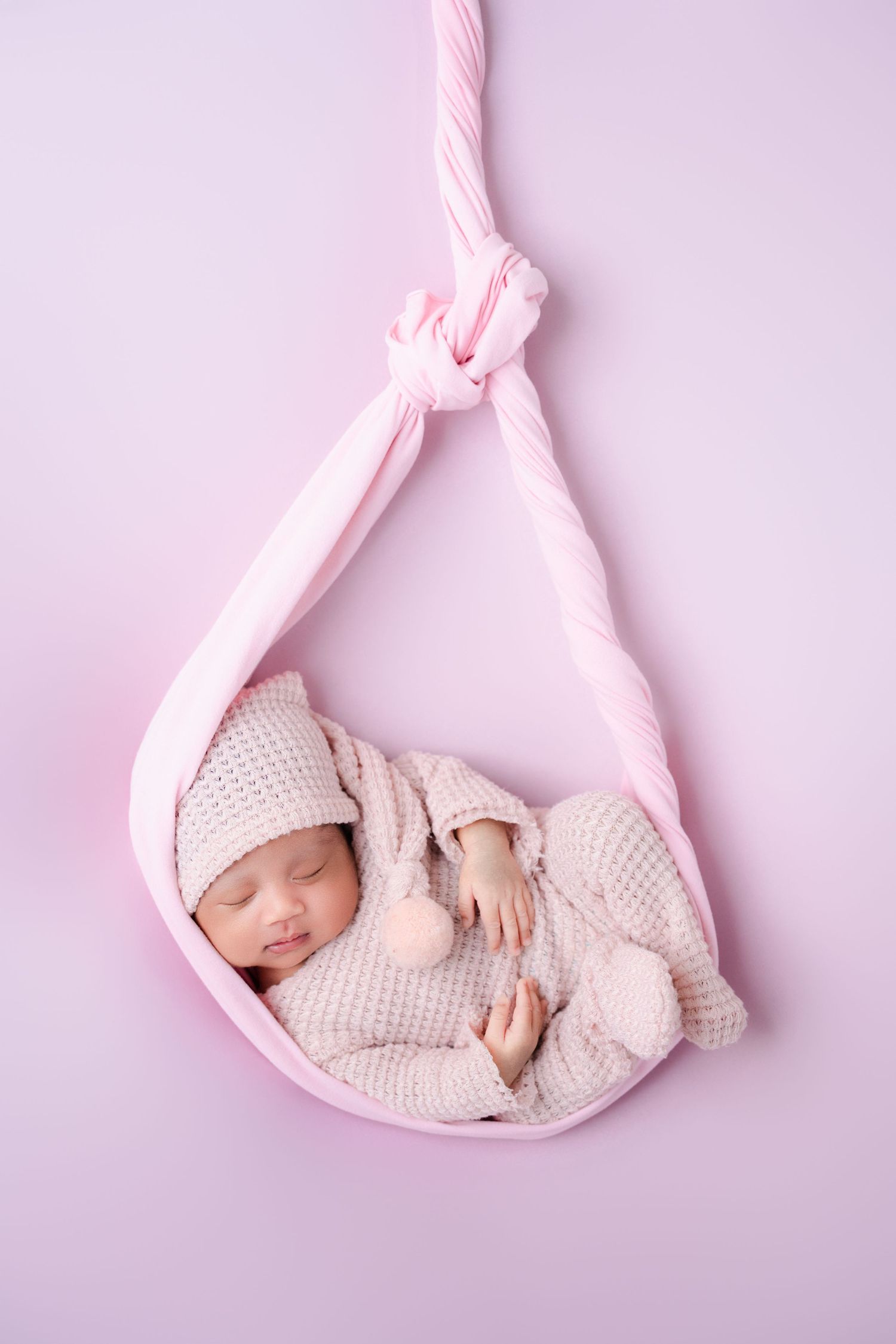 Sleeping newborn in knit outfit resting in pink fabric hammock on lilac background.