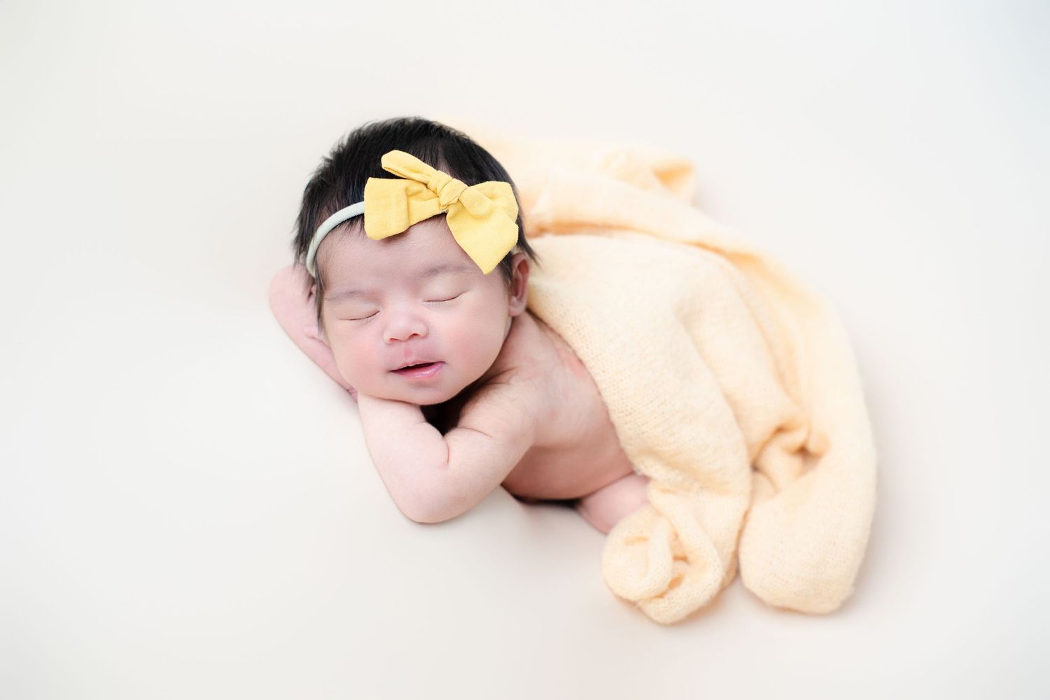 Sleeping newborn with yellow bow accessory wrapped in peach blanket.
