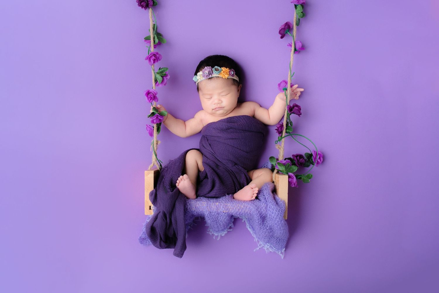 Baby wrapped in purple fabric sleeping on rustic wooden swing with purple flowers.
