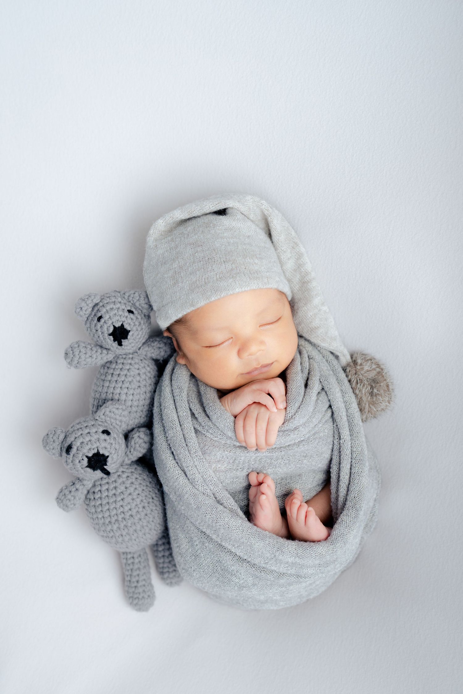 Sleeping newborn wrapped in soft gray fabric lying next to knitted teddy bear in minimalist photo series.