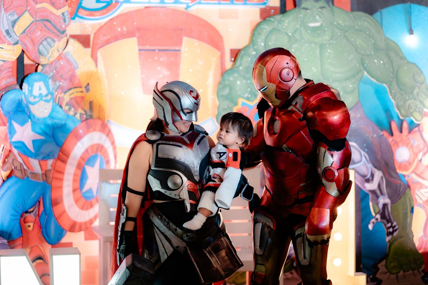 Child poses with Thor and Iron Man costumed characters in front of colorful Marvel Avengers themed backdrop.