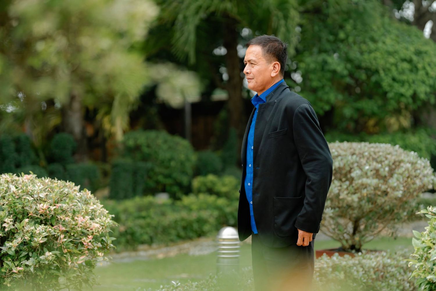 Middle-aged Asian man in black jacket and blue shirt strolling through a lush green garden.