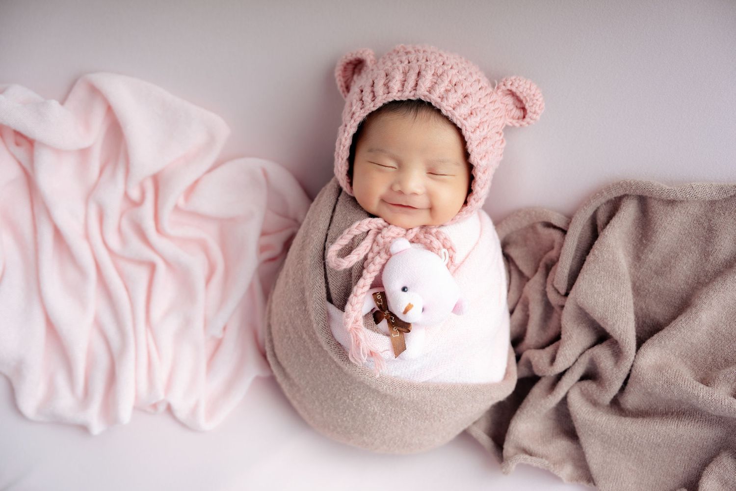 A sleeping newborn baby wearing a pink knit hat lies peacefully on soft gray and pink blankets.
