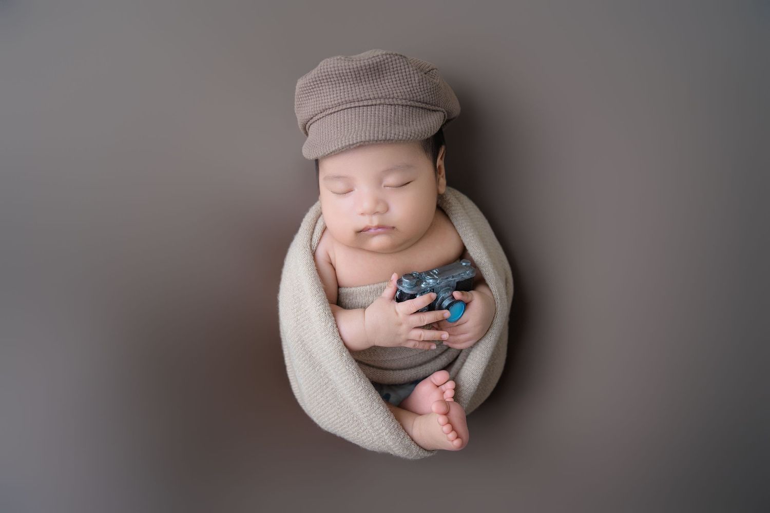 Sleeping newborn wearing gray newsboy cap swaddled in beige wrap against neutral background.