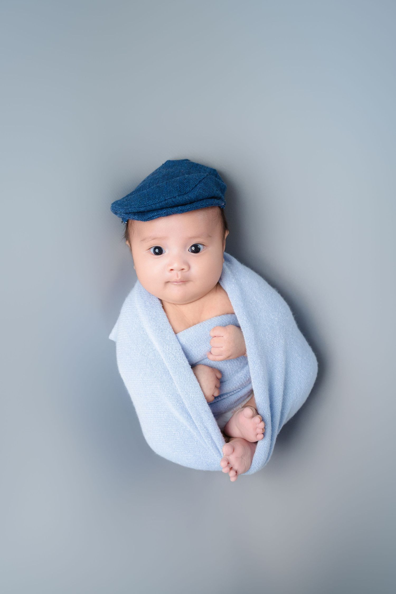 Baby wearing blue newsboy cap and wrapped in light blue blanket against gray backdrop.