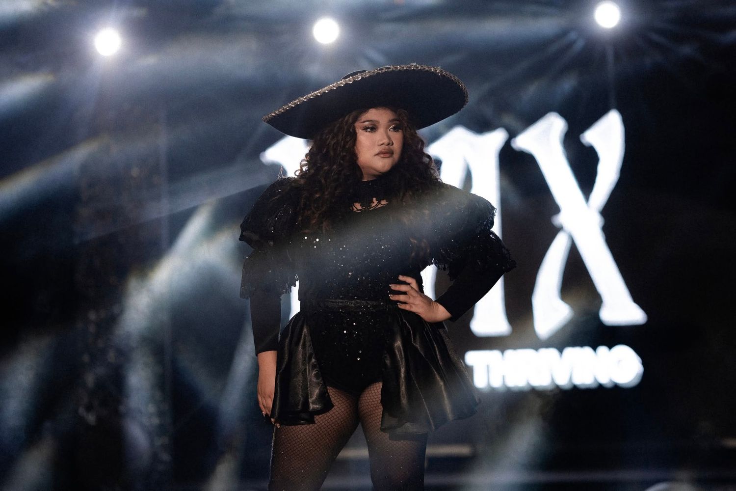 Woman in black outfit and sombrero poses on a dramatic stage with spotlights at a music awards show.