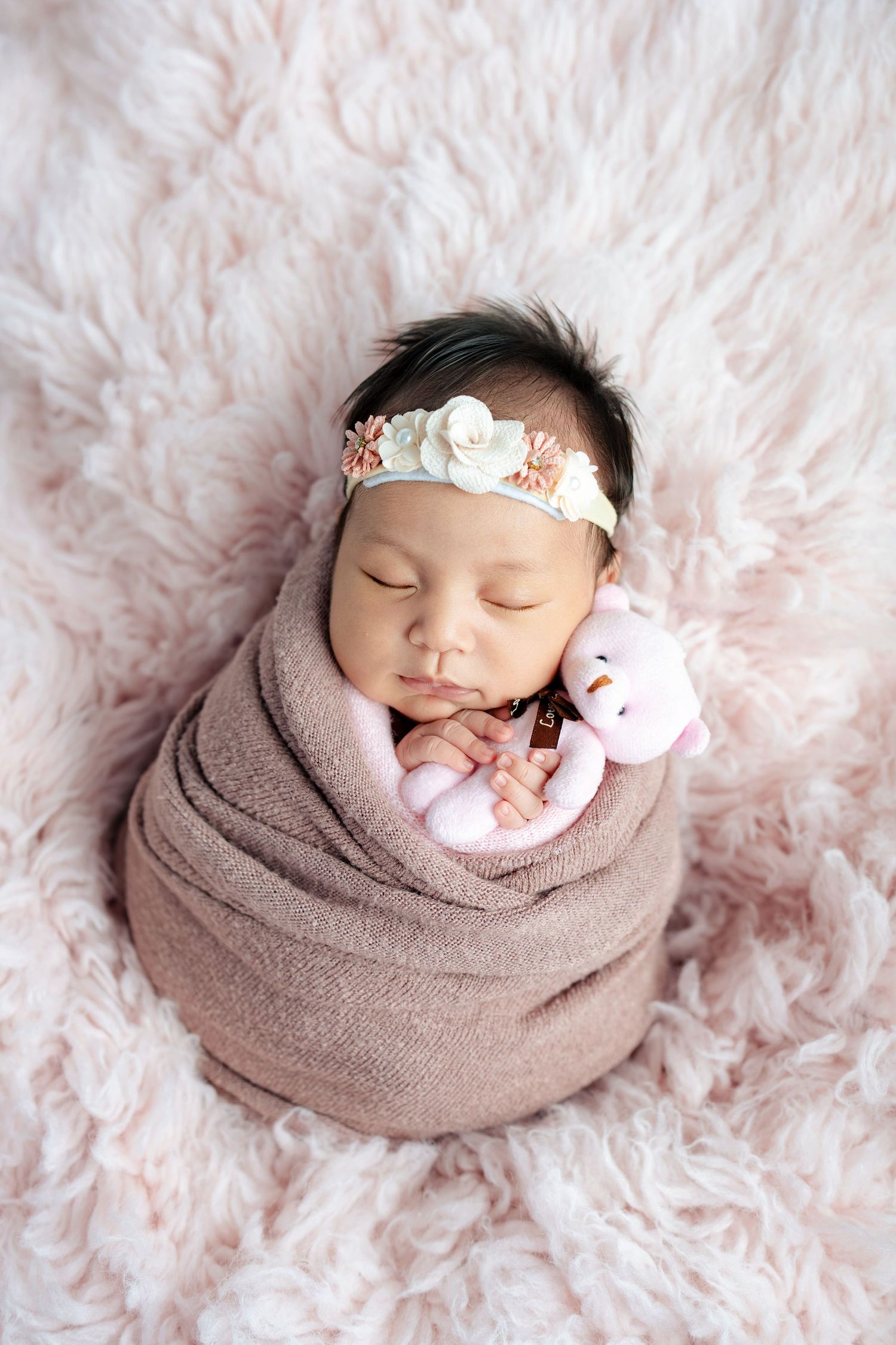 Newborn baby peacefully sleeping on fluffy white rug while wrapped in soft brown swaddle with floral headband.