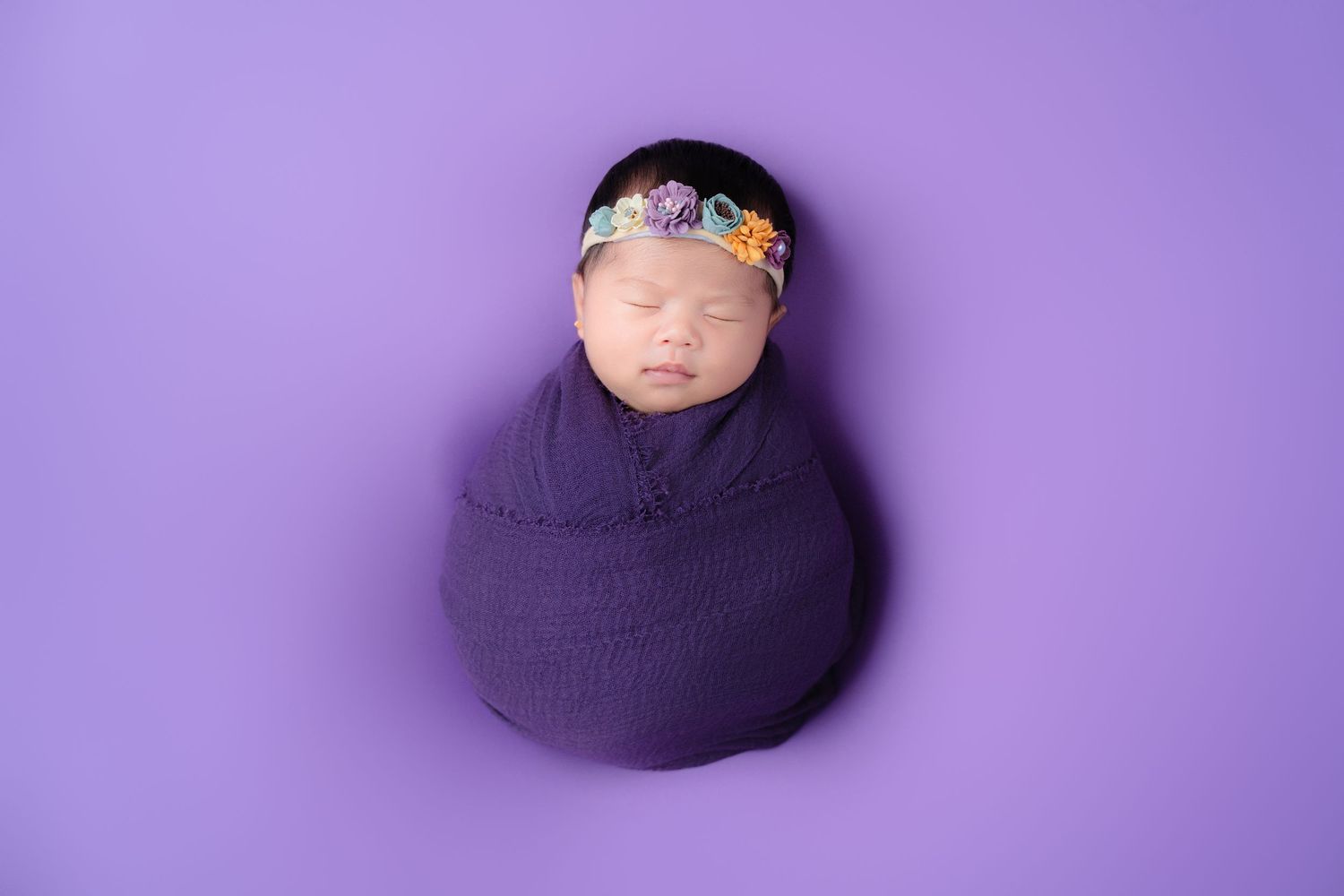 Sleeping newborn wrapped in purple fabric with floral headband on purple backdrop.