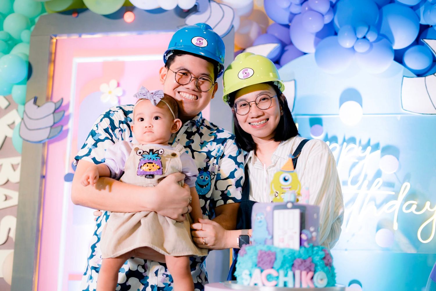 Family of three celebrating a baby's first birthday party with colorful balloon decorations and matching outfits.