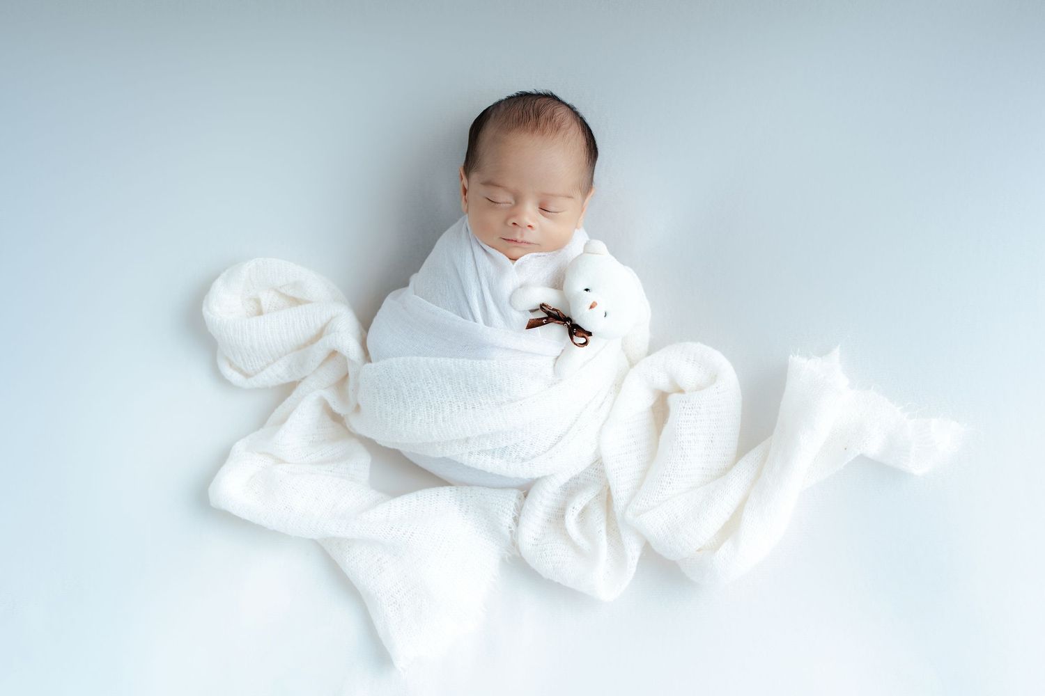 Sleeping newborn baby wrapped in soft white blanket on light blue background.