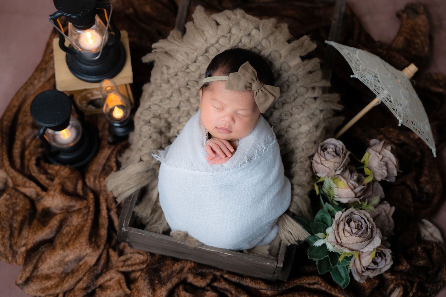 Peaceful baby wrapped in white swaddle with decorative lanterns and roses on brown fabric.
