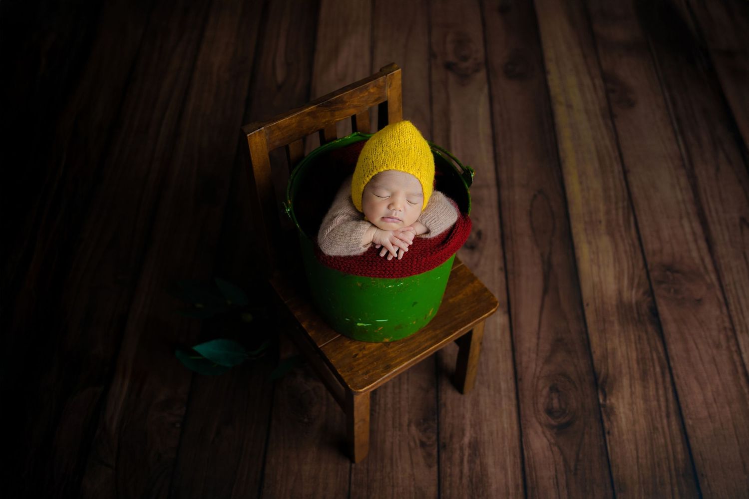 Sleeping newborn in yellow knit wrap nestled in green bucket on wooden chair.