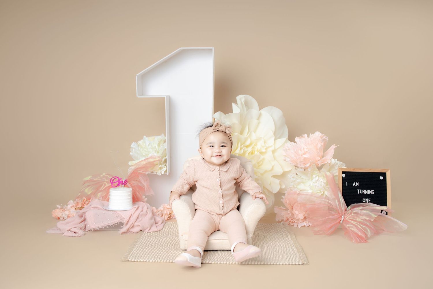 Baby in pink tutu and pearls poses on beige backdrop with soft pink floral decorations.