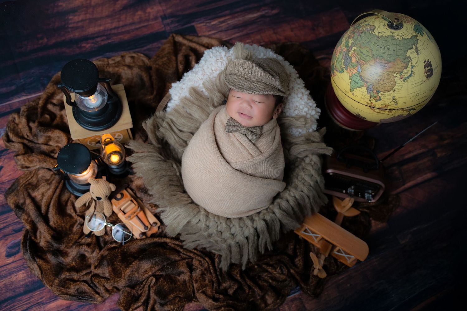 Vintage-styled newborn photo with globe, wooden toys and swaddled baby on textured fabric.