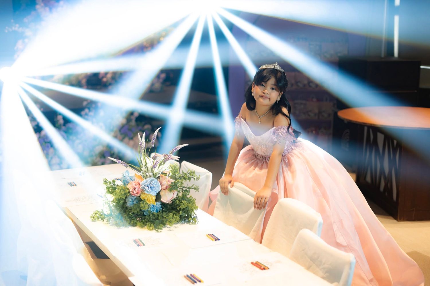 Young woman in elegant pink ball gown posing beside floral centerpiece under dramatic stage lighting.