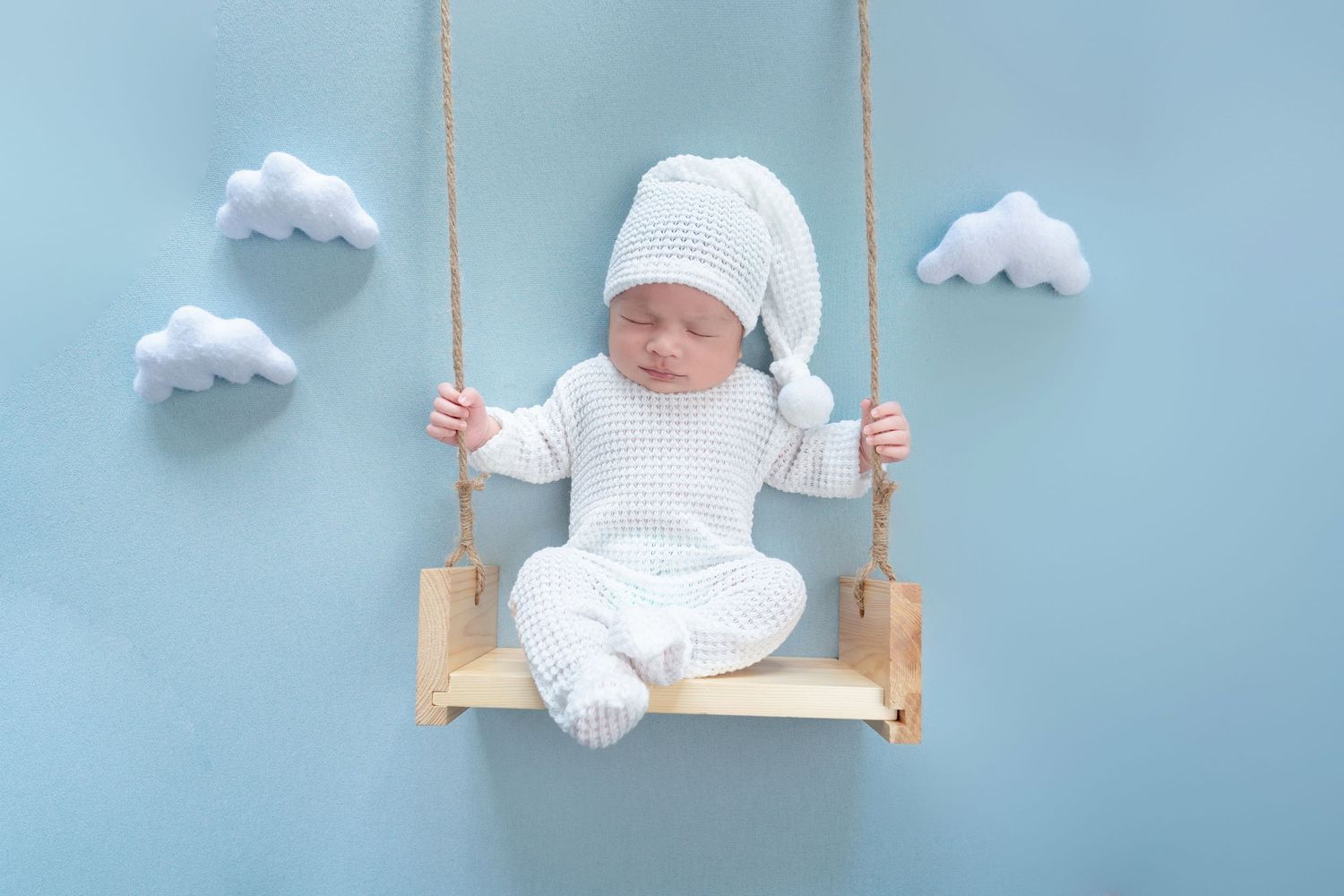 Baby in white outfit on wooden swing with cloud decorations against blue background.