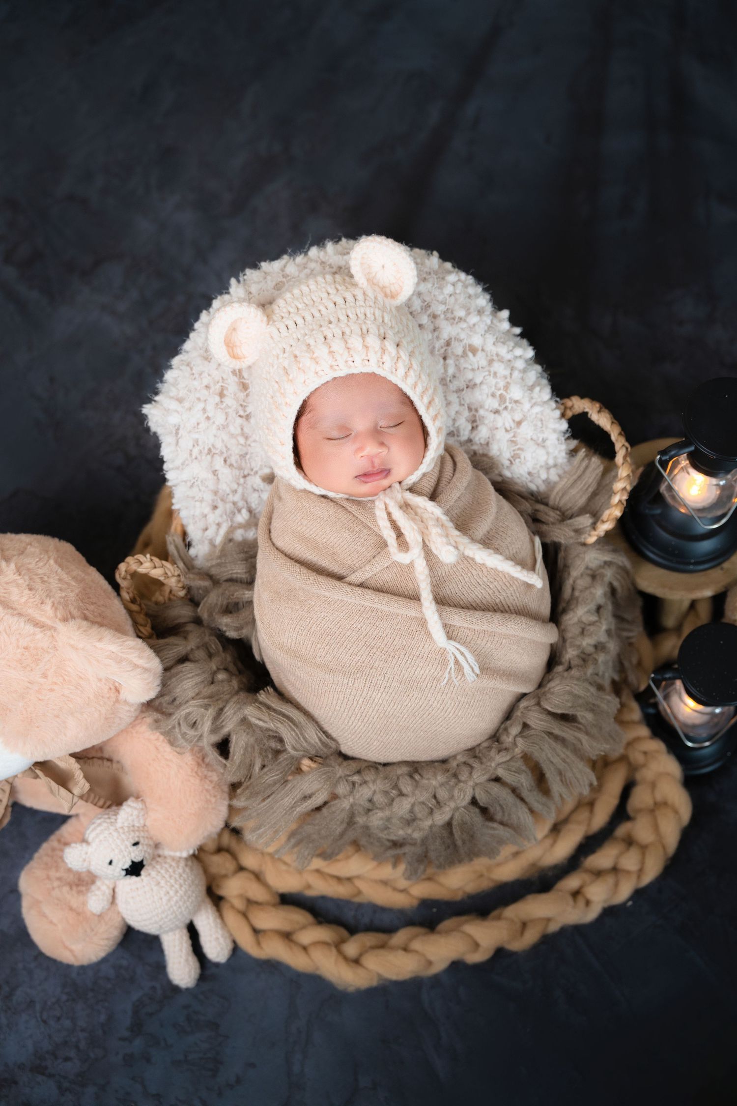 Artistic newborn portrait with fluffy white bonnet and beige wrap surrounded by plush toys and lanterns.