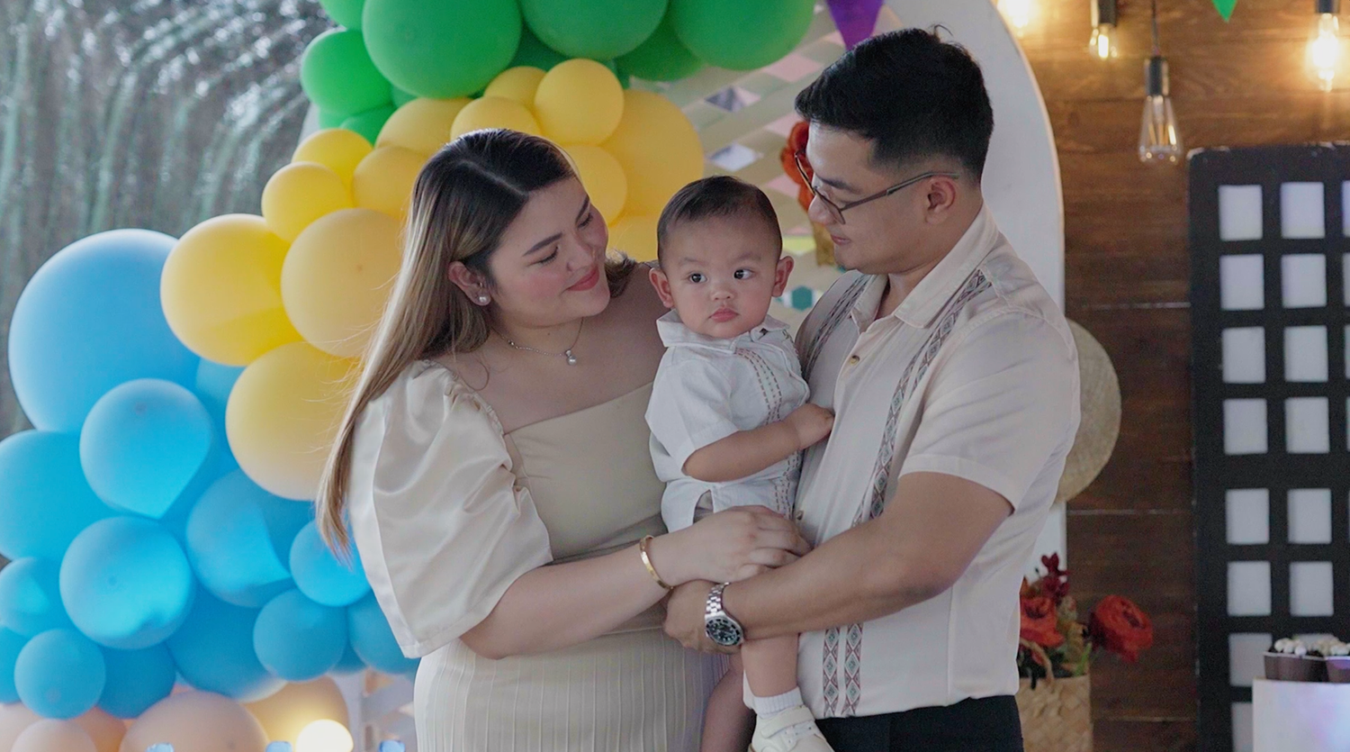 A young Asian family with a baby boy poses in front of colorful balloons at a birthday celebration.