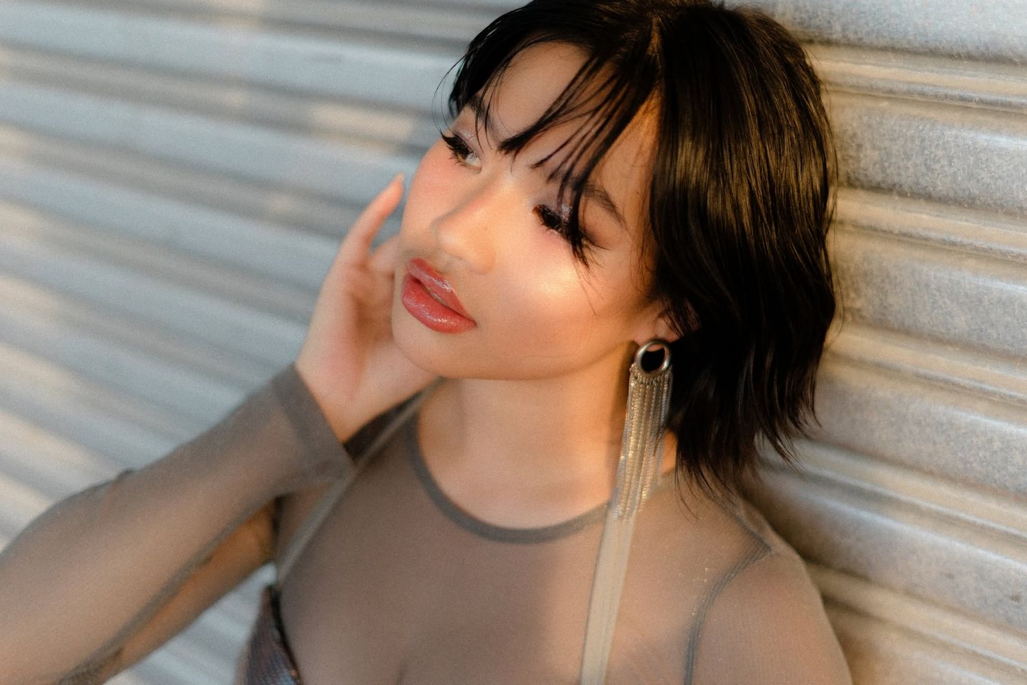 Young woman with short dark hair and long earrings poses against a corrugated metal wall in a sheer top.