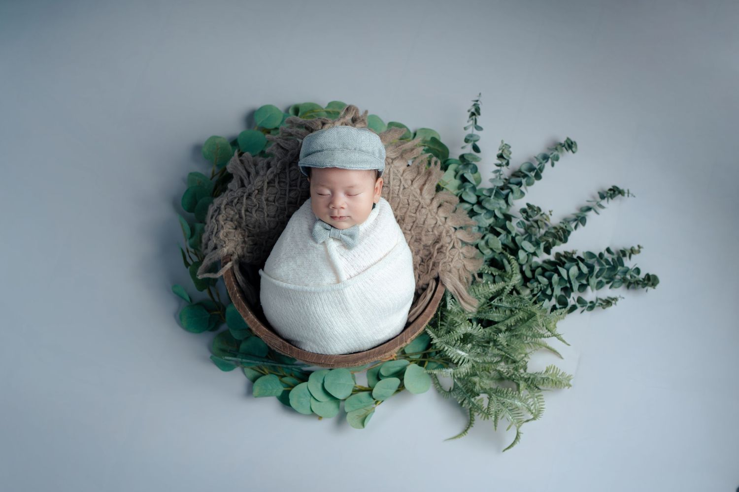 Sleeping newborn baby wrapped in soft gray swaddle with delicate green foliage decorative elements.