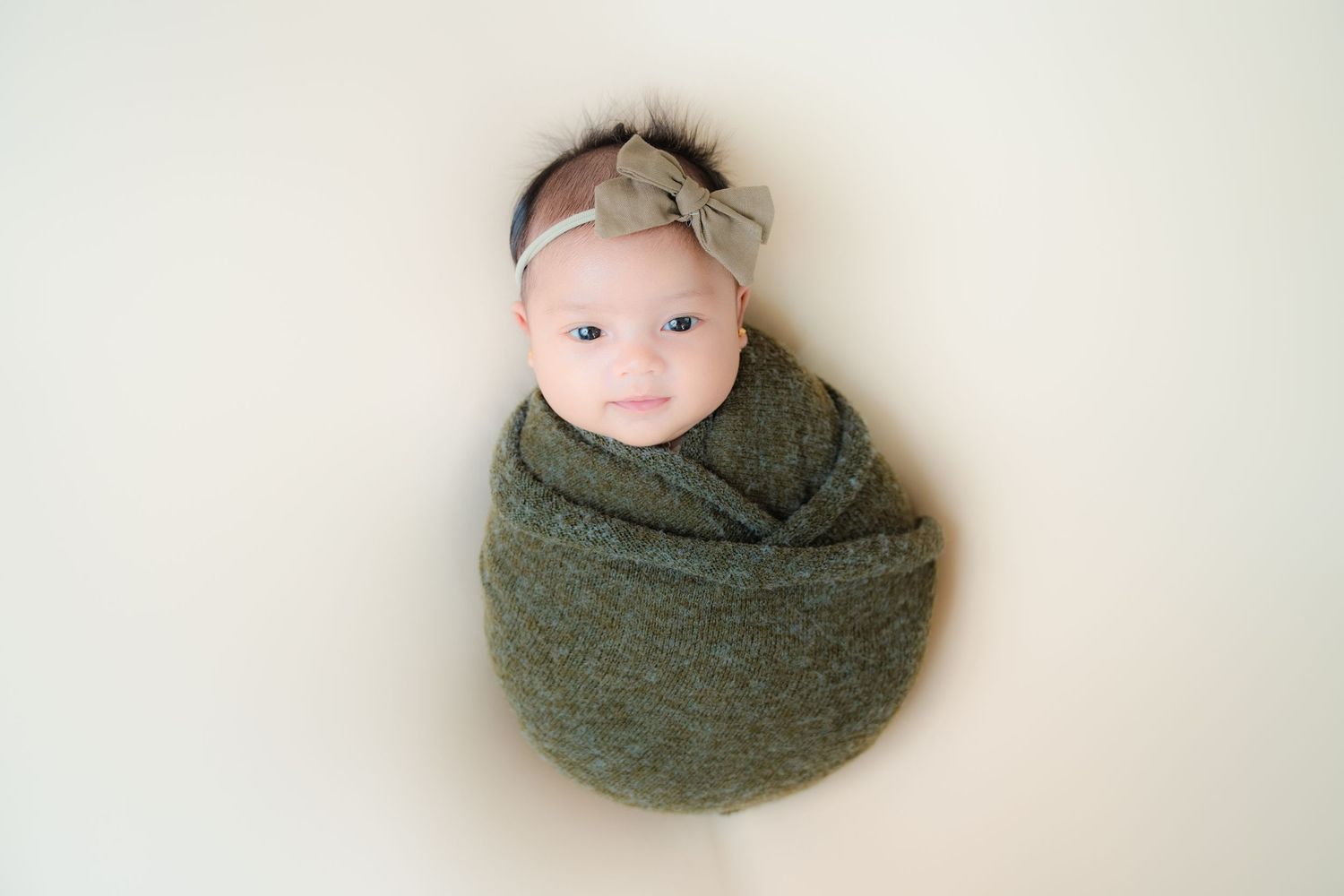Newborn swaddled in olive green wrap with headband on light background.