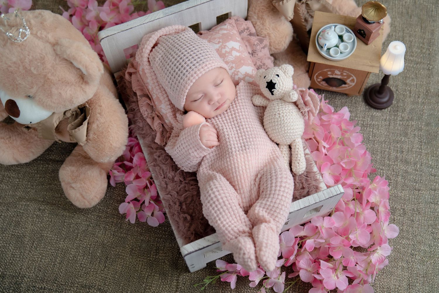 Baby in pink knit outfit sleeping in white crate surrounded by pink flowers and plush toys.