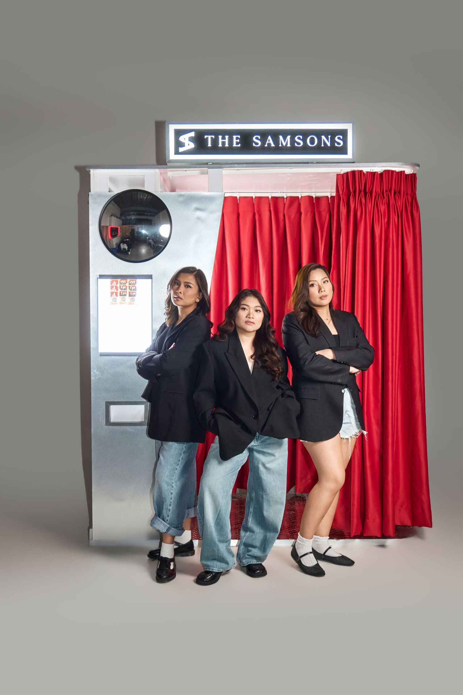 Three women in black blazers pose confidently in front of The Samsons photo booth with red curtains.