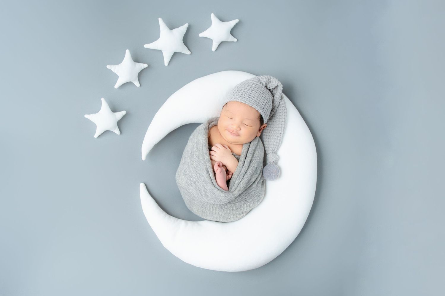 Sleeping baby wrapped in grey swaddle posed on decorative white crescent moon with stars.