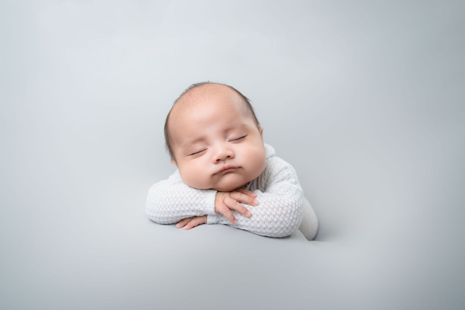 Sleeping newborn in white wrap posed on soft gray background during professional photoshoot.