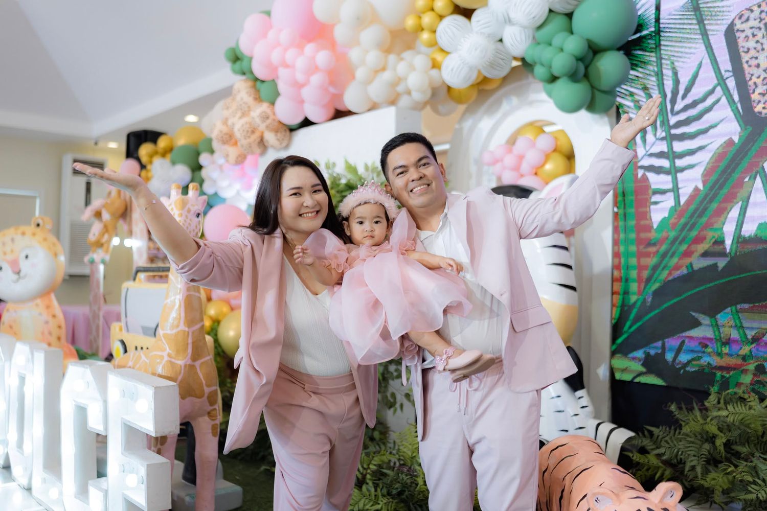Family of three in pink outfits celebrating a jungle-themed birthday party with colorful balloon arch backdrop.