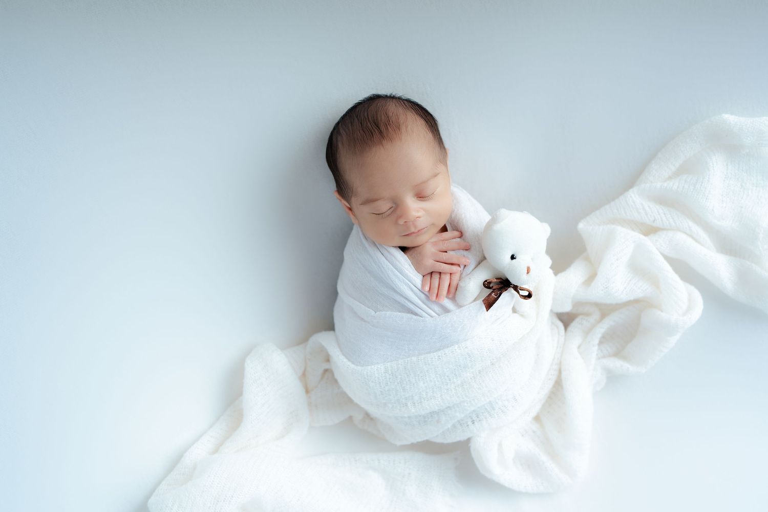 A peaceful newborn baby sleeps wrapped in soft white blankets against a light background.