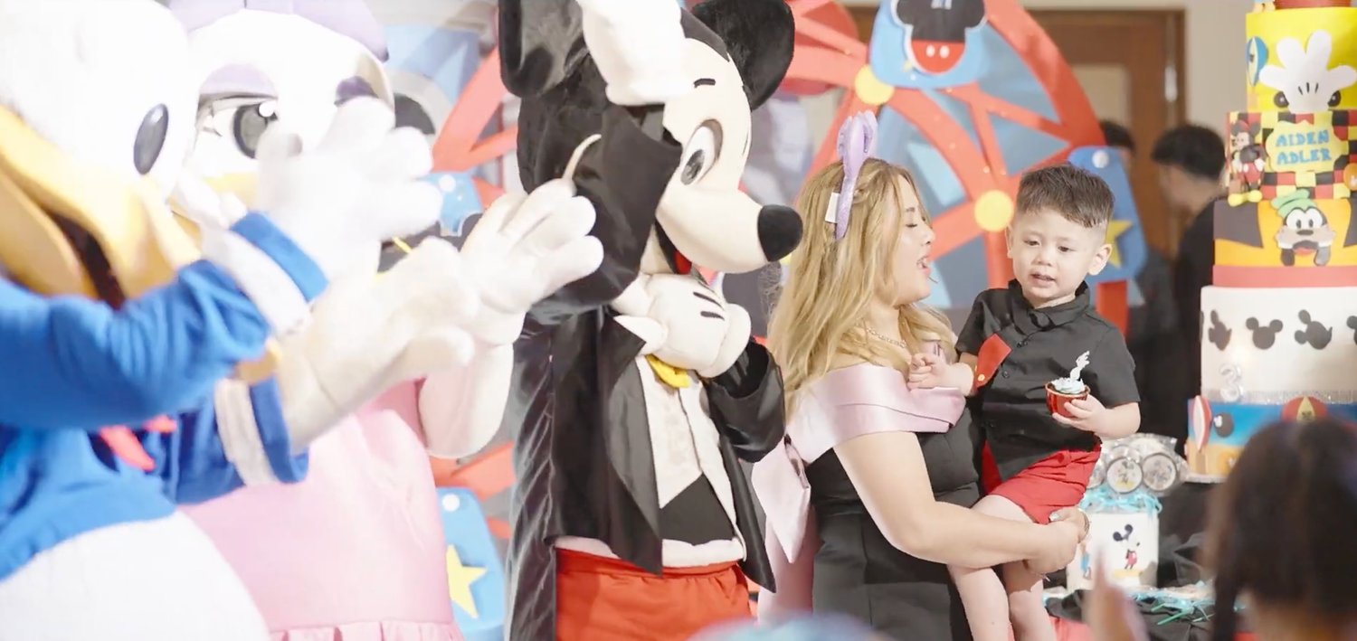 A woman holds a toddler near Mickey Mouse and Donald Duck mascots at a colorful Disney-themed birthday party.