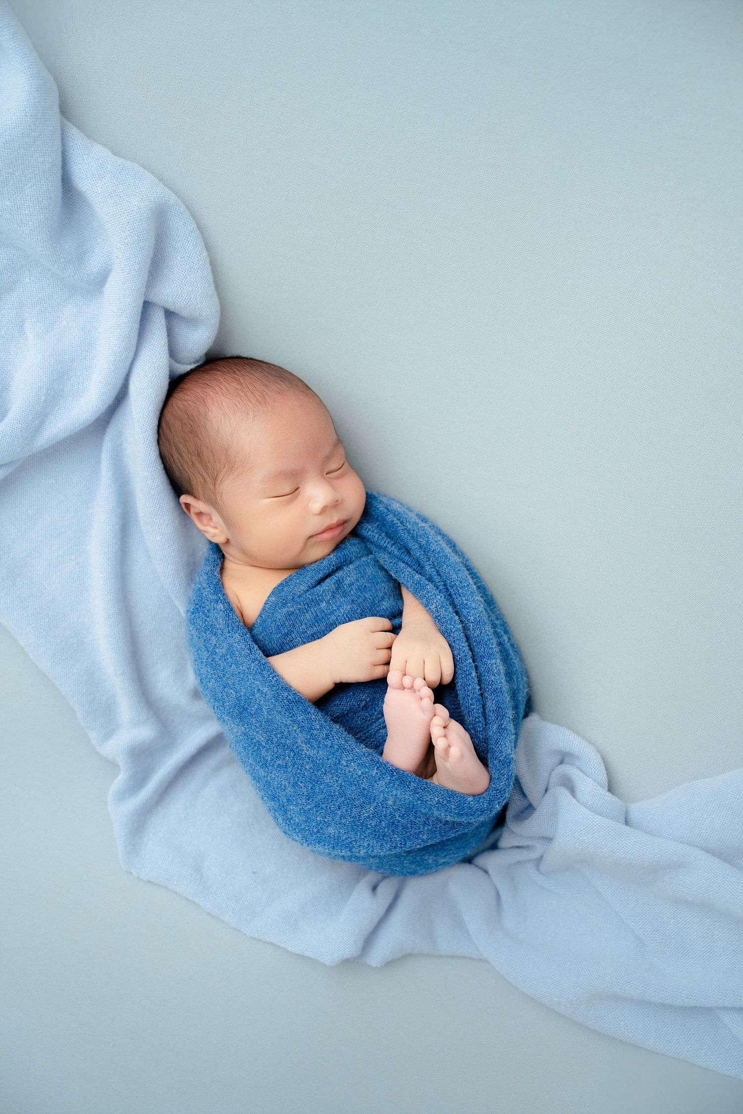 Sleeping newborn wrapped in blue fabric on flowing light blue textile backdrop.
