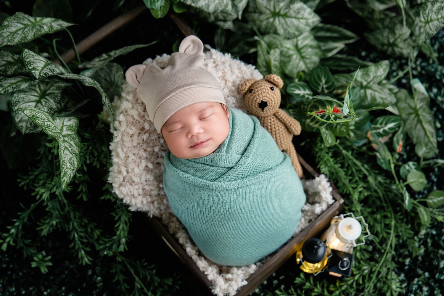 Sleeping baby in mint wrap with teddy bear among greenery and vintage props.
