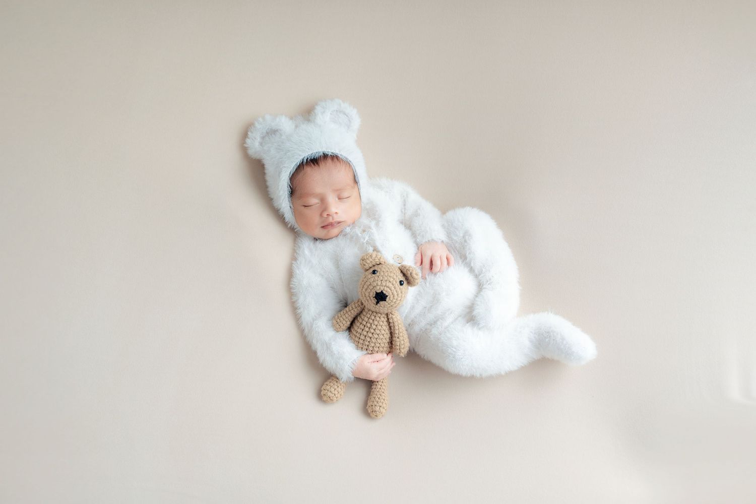 A newborn baby in white sleepwear rests peacefully on a soft white blanket during a professional photo session.