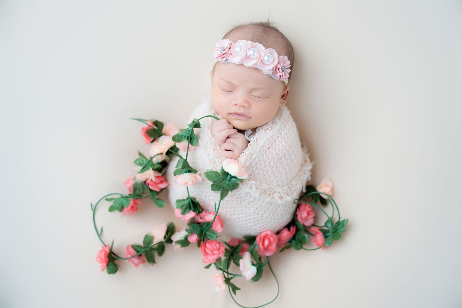 Newborn wrapped in white swaddle with floral headband and pink rose vine decorations.