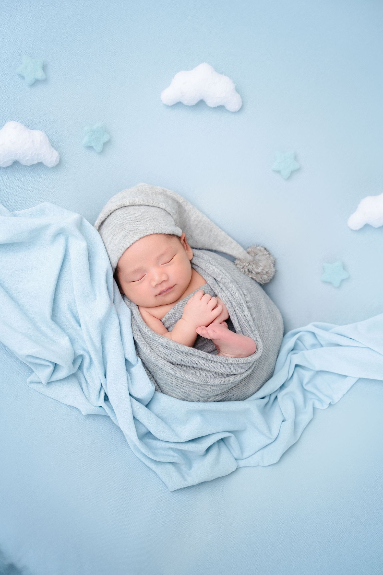 Newborn photography setup with soft blue fabrics and decorative white clouds against a light blue backdrop.