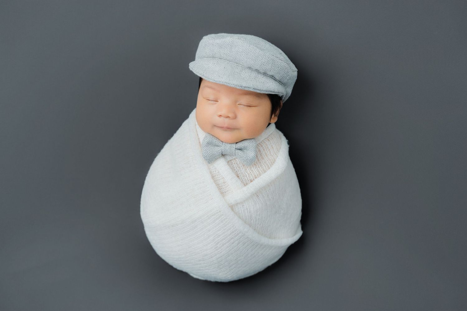 Sleeping infant wrapped in soft gray fabric against dark background.