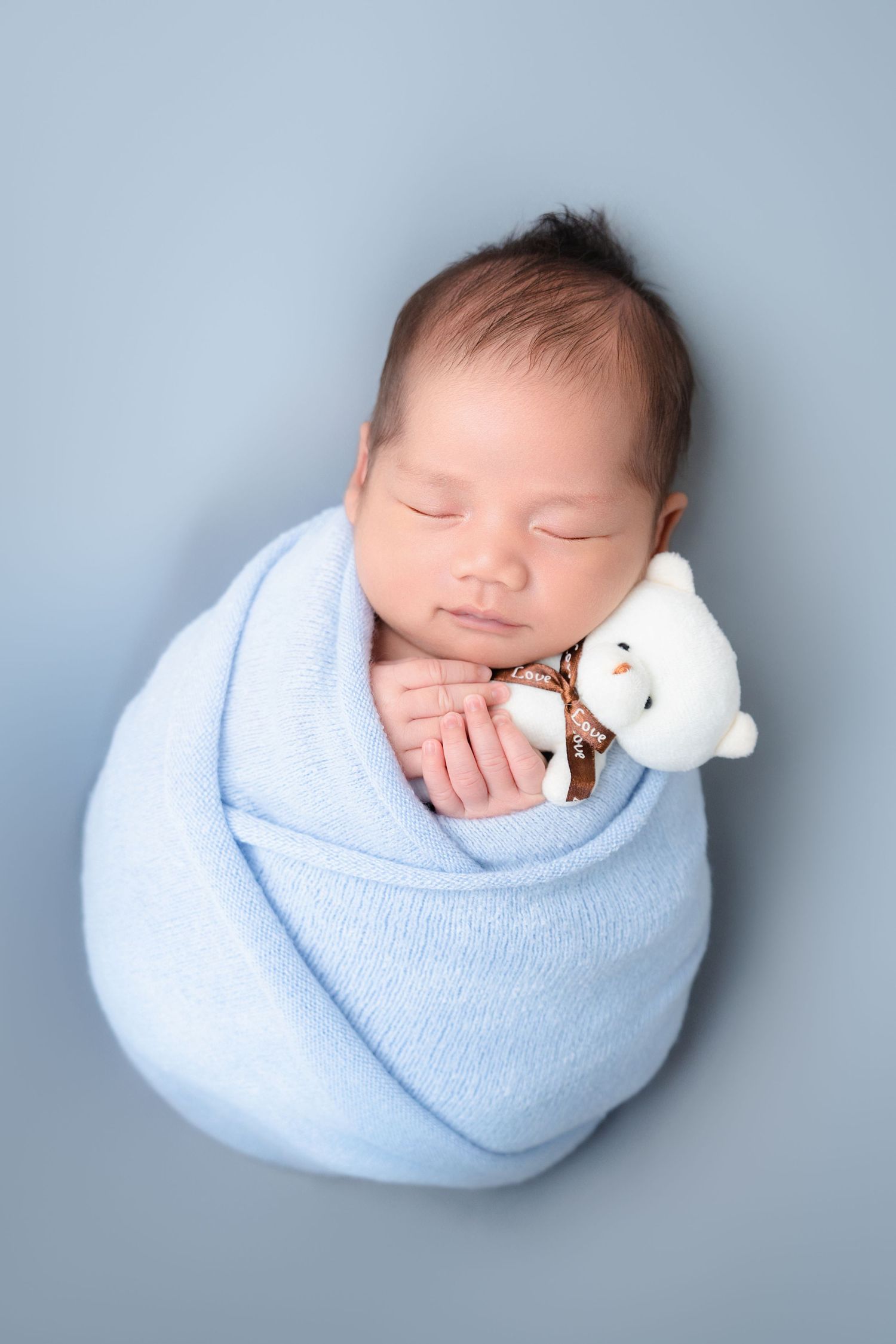 Sleeping newborn wrapped in light blue blanket holding small white teddy bear.