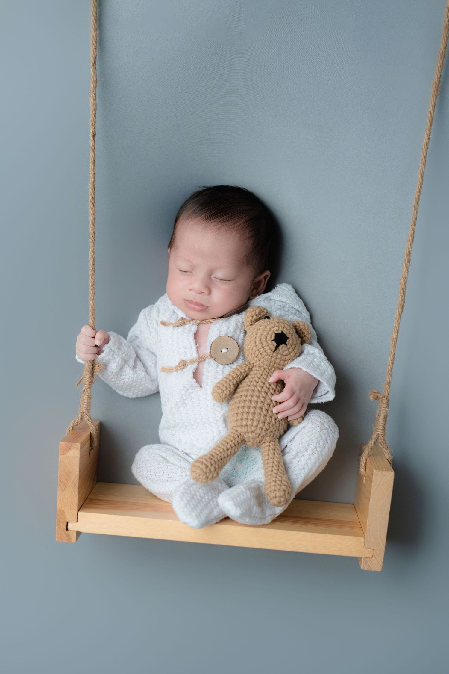 Sleeping baby in white outfit with teddy bear on wooden swing against grey backdrop.