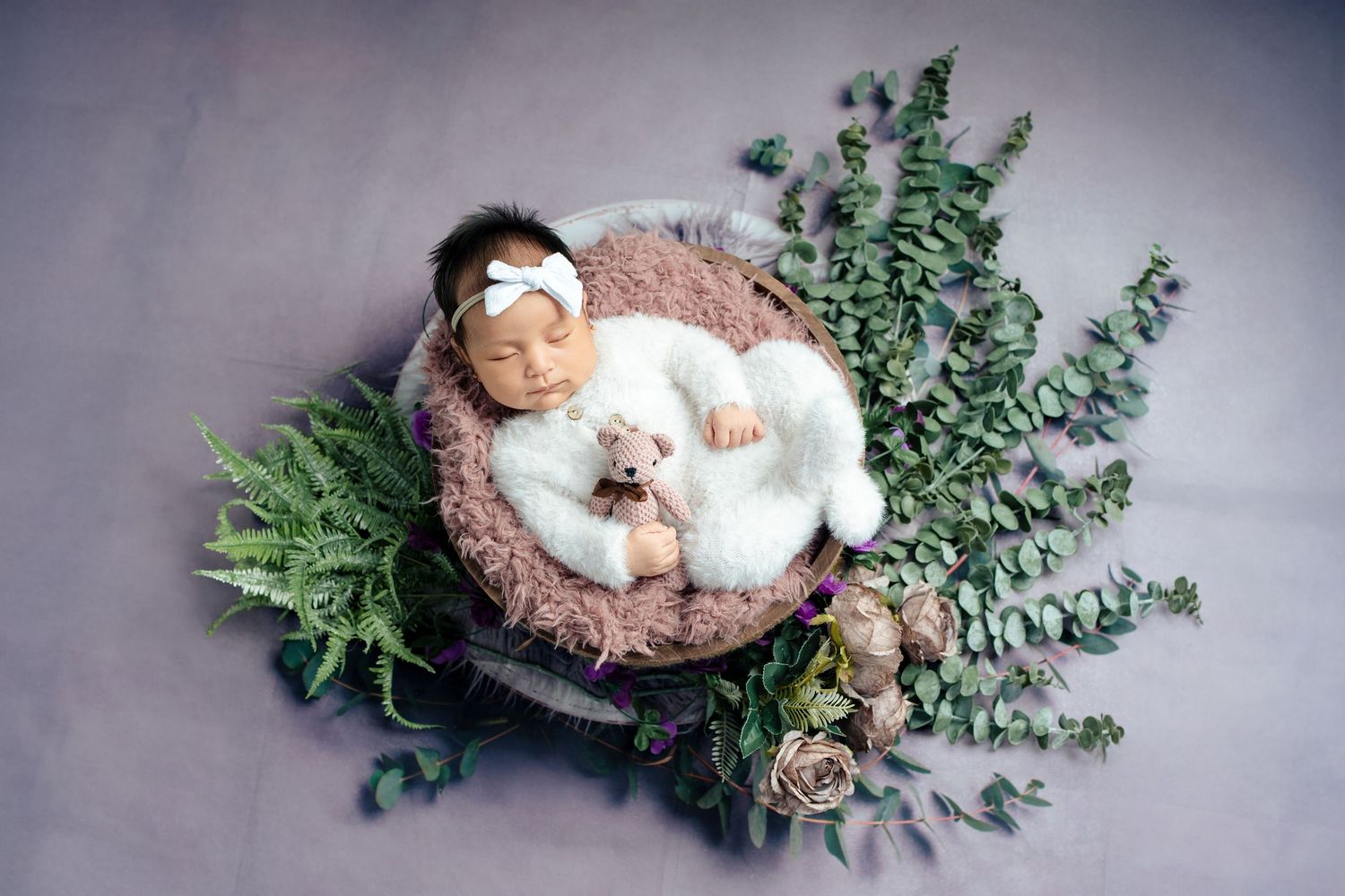 Sleeping newborn wrapped in brown fabric nestled in a rustic wooden bowl with green eucalyptus branches.