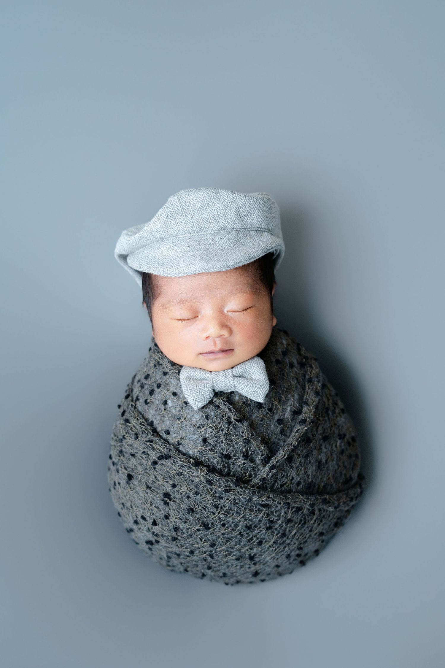 Newborn baby wearing grey flat cap and bowtie wrapped in textured grey blanket on light background.
