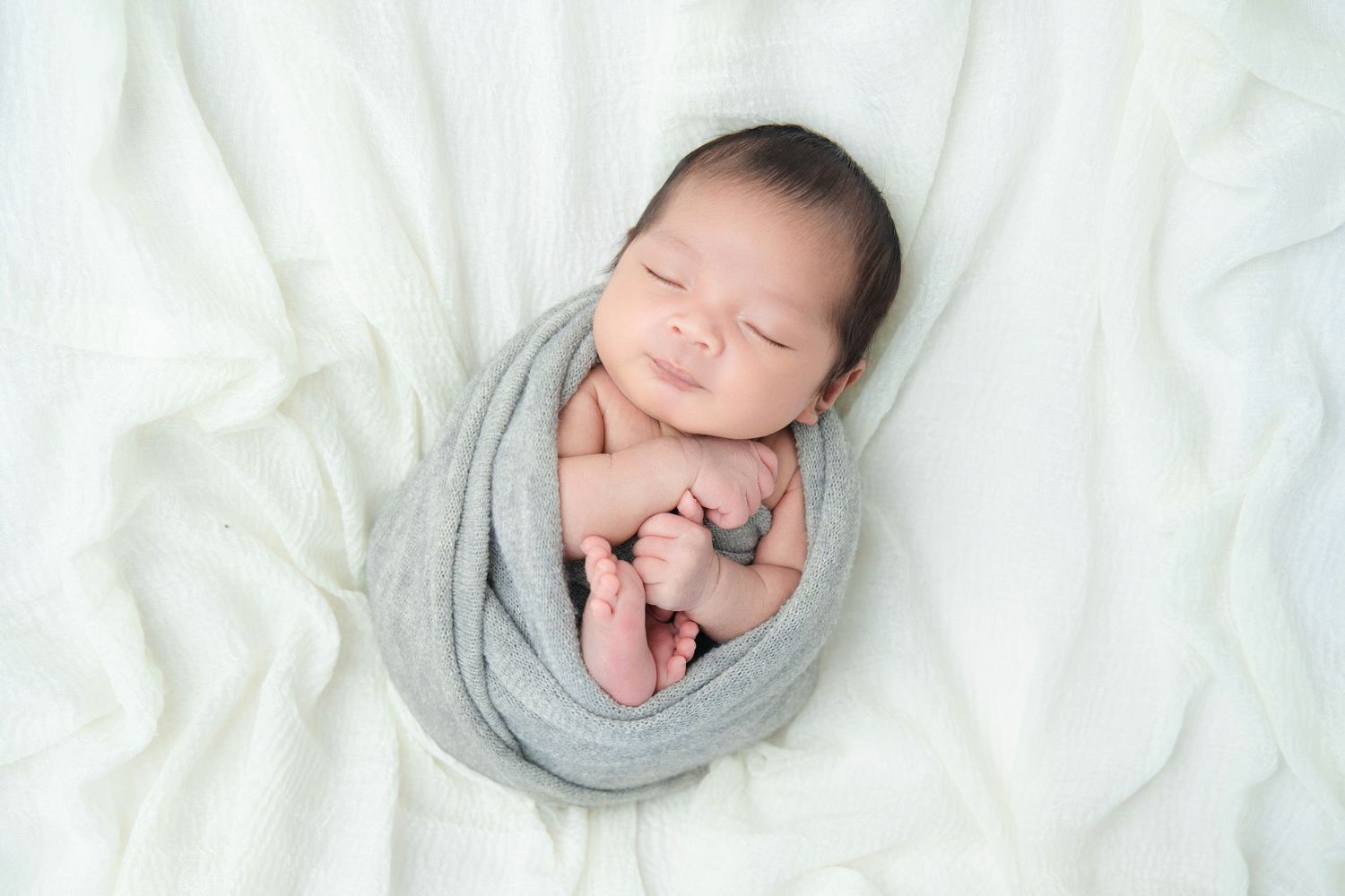 Sleeping newborn wrapped in soft gray fabric on white textured background.