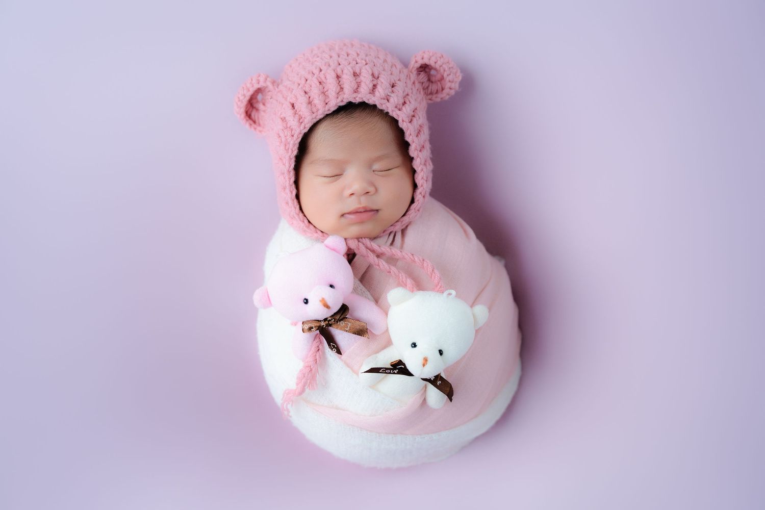 Sleeping newborn in pink knit bear bonnet with plush toys on purple backdrop.